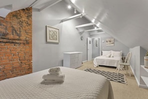 A bedroom with a brick wall, a bed with white bedding, a bedside table, a chair, and a small desk with a chair.
