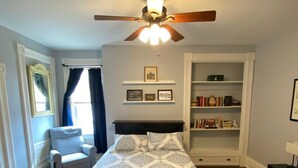 Un dormitorio con cama, una silla, ventilador de techo, estantería y ventana con cortinas.