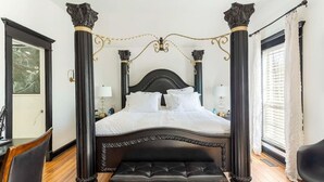 A four-poster bed with a dark headboard and footboard, flanked by ornate columns, in a room with a wooden floor and a window with white curtains. - Pool + Hot Tub + Theater · Countryside Retreat (Hermitage)