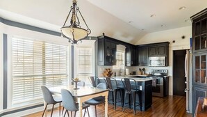 A modern kitchen with dark cabinetry, a central island, and a dining area with a table and chairs. - Pool + Hot Tub + Theater · Countryside Retreat (Hermitage)