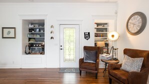 A living room with a brown leather sofa, a matching armchair, a wooden floor, a white door with a glass panel, and a bookshelf with various items. - Pool + Hot Tub + Theater · Countryside Retreat (Hermitage)