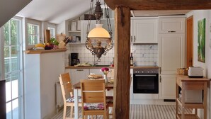 Een keuken met witte kasten, een houten eettafel met stoelen, een hangende lamp en een raam met uitzicht op groen.
