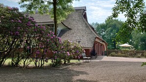 Een traditioneel huis met rieten dak, omgeven door een grindpad en bloeiende struiken.