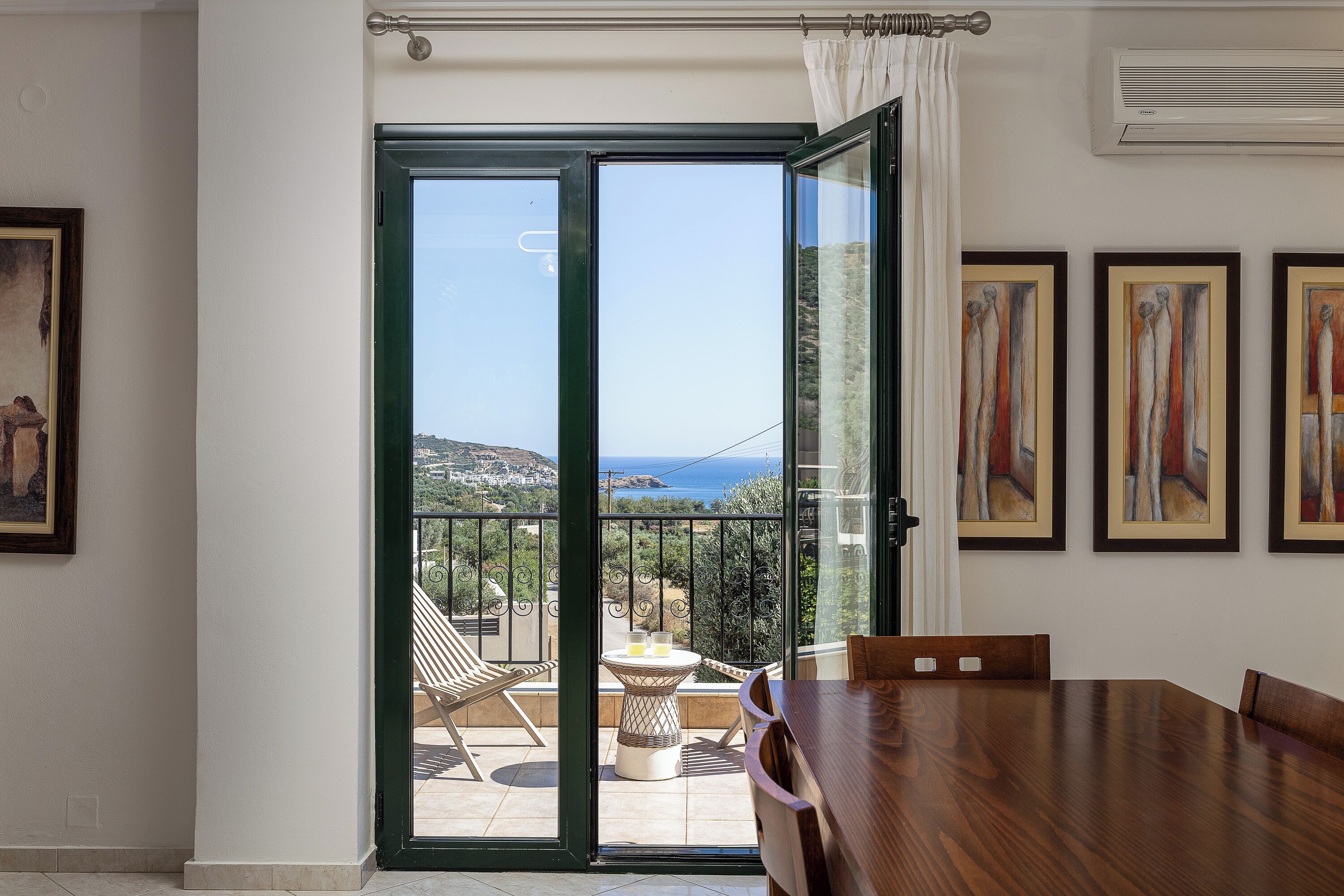 Une pièce avec une table à manger en bois, un balcon donnant sur la mer et des œuvres d’art encadrées accrochées au mur.