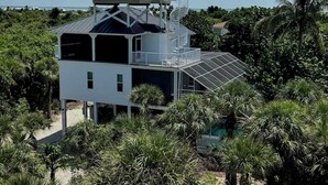 A modern two-story house with a glass balcony and a large window, surrounded by lush greenery. - Manatee Haven: Screened Pool, Spa, Elevator (Captiva)