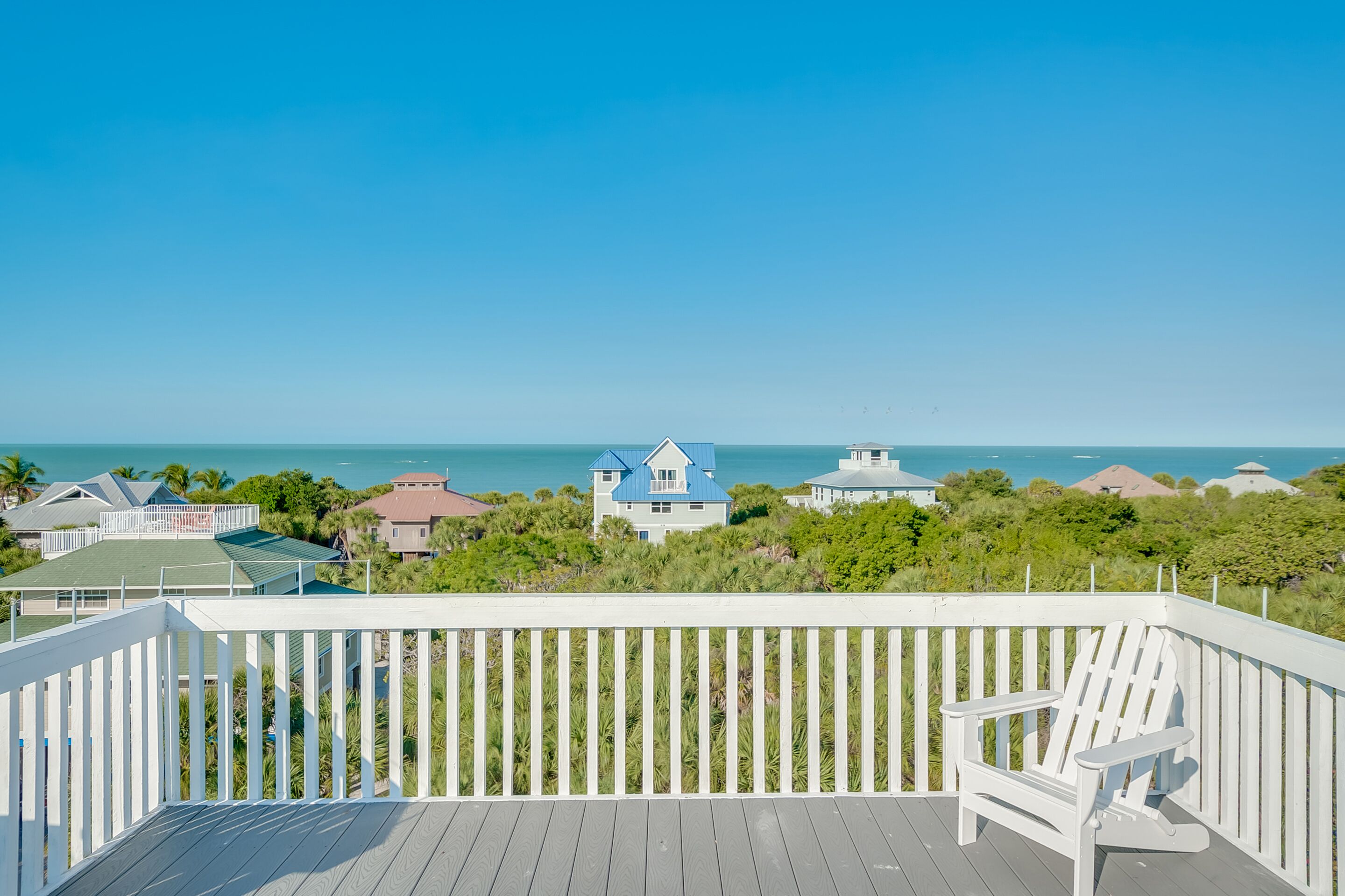 A wooden deck with a white railing, a white Adirondack chair, and a view of the ocean and houses in the distance.