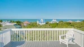 A wooden deck with a white railing, a white Adirondack chair, and a view of the ocean and houses in the distance. - Manatee Haven: Screened Pool, Spa, Elevator (Captiva)