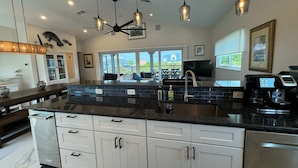 A modern kitchen with a large island, white cabinets, and a black countertop. There are pendant lights, a ceiling fan, and a view of the outdoors. - Manatee Haven: Screened Pool, Spa, Elevator (Captiva)