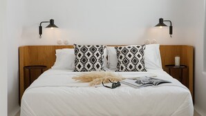 A neatly made bed with white bedding, two patterned pillows, a fluffy decorative item, and a magazine. There are two bedside tables with lamps, and a small round table with a candle. - Alio Suites-Suite N2 with private swimming pool (Νάξος)