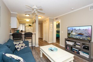 A living room with a sofa, chairs, a coffee table, a TV, and a kitchen in the background.
