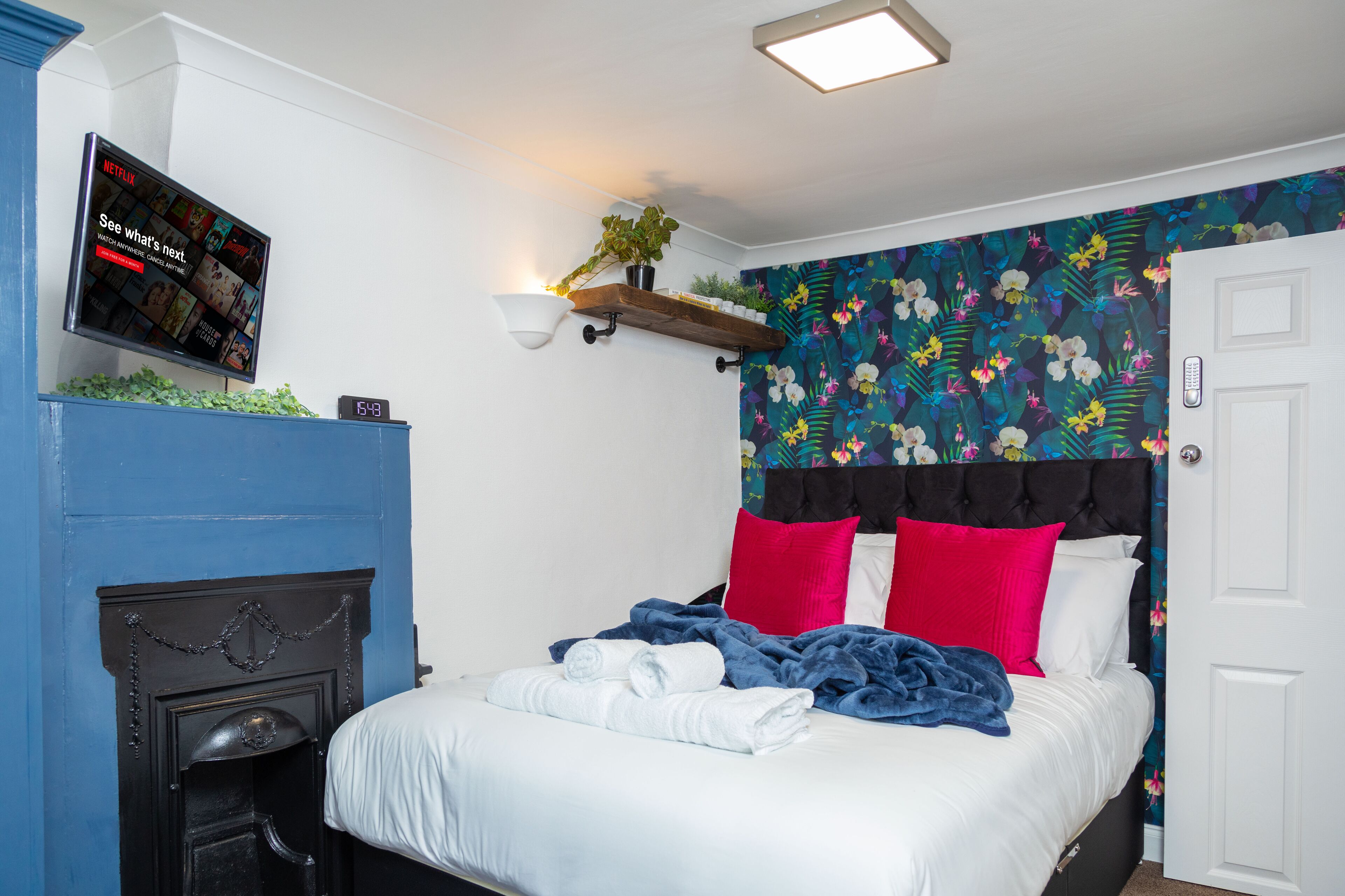 En pænt redt seng med hvide og blå sengetæger, et monteret fjernsyn der viser Netflix, en vægmonteret hylde med en plante, og tapet med blomstermønster.