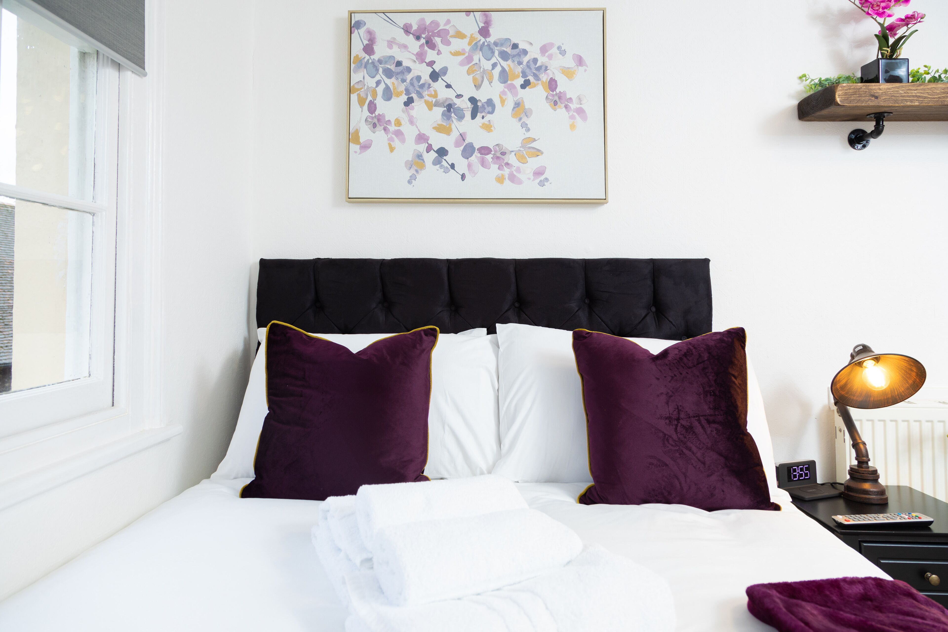 A neatly made bed with white linens and purple pillows, a bedside table with a lamp, and a wall-mounted shelf with a plant.