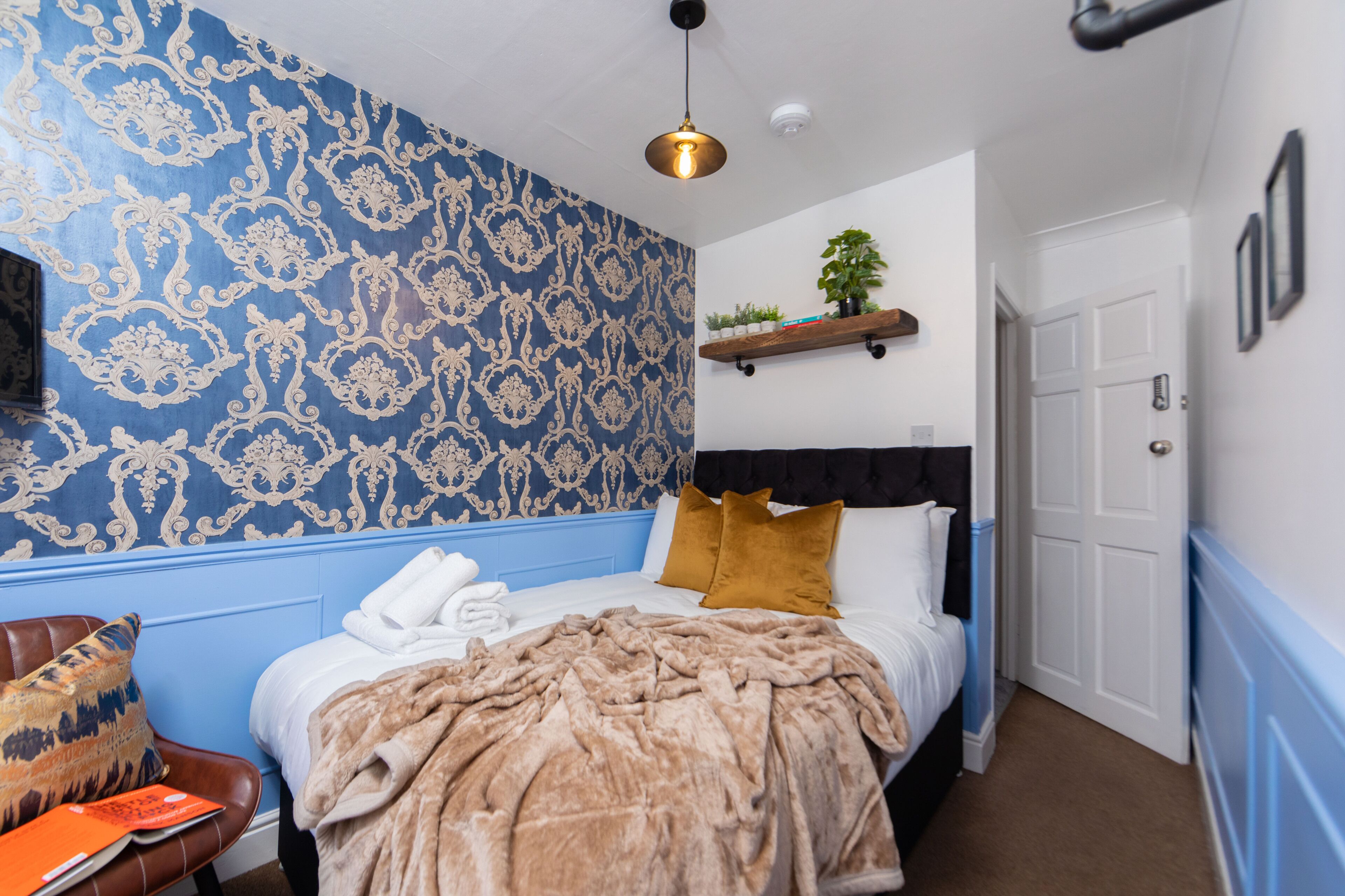 A bedroom with a patterned wallpaper, a bed with a brown blanket, a wooden shelf with plants, and a door.