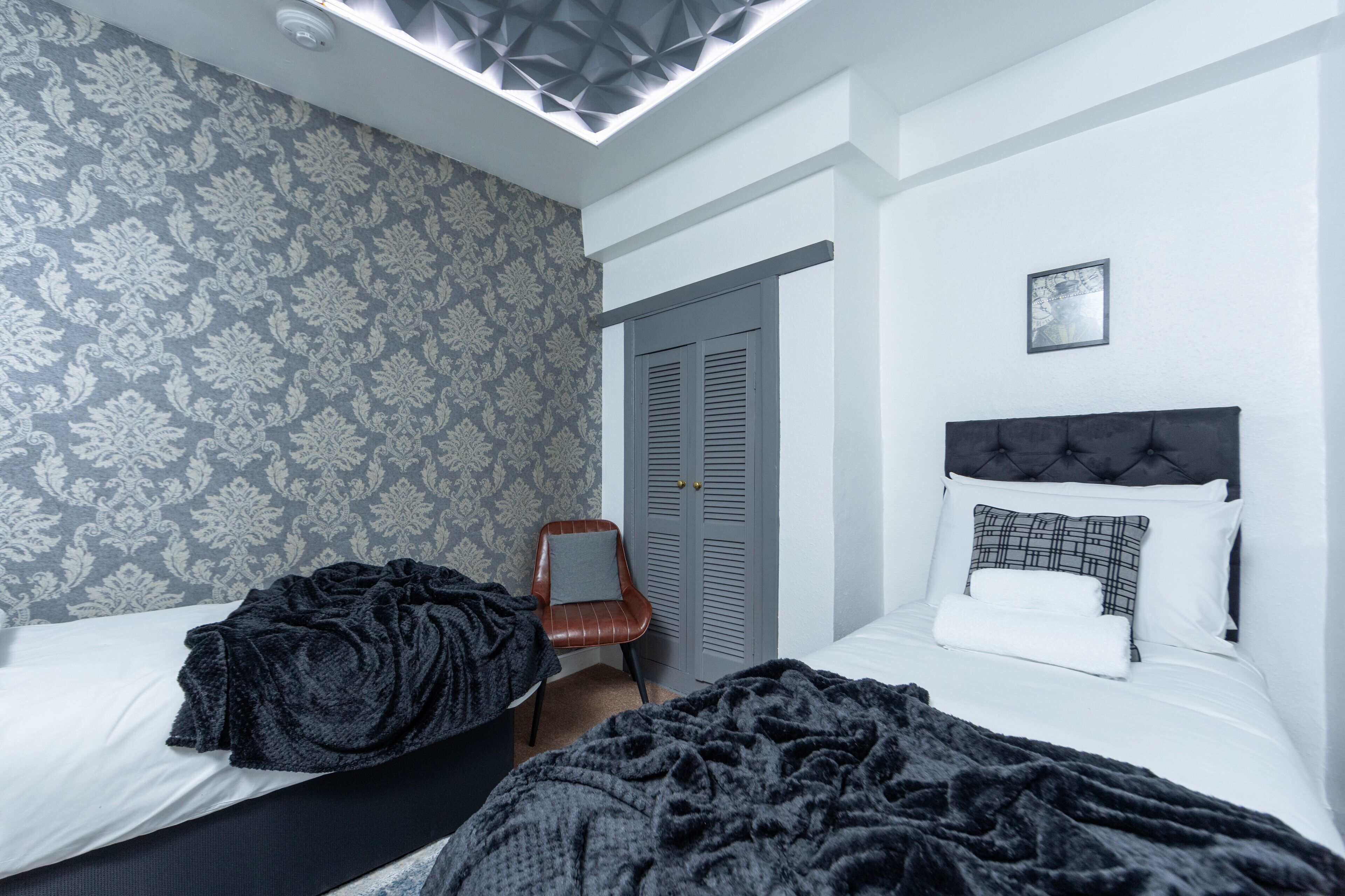 Un dormitorio con dos camas, un papel tapiz estampado, una lámpara en el techo, una puerta y un cuadro enmarcado en la pared.