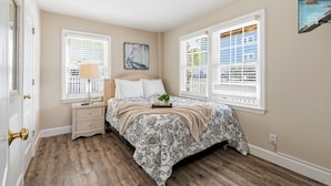 Une chambre à coucher avec un lit, deux fenêtres équipées de stores, une table de chevet, une lampe, un tableau et une vue sur l’extérieur.