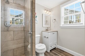 A bathroom with a glass shower enclosure, a toilet, a sink with a vanity, and a window with blinds.
