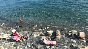 Ein felsiger Strand mit klarem Wasser, mehreren Liegestühlen und einer Person, die im Wasser steht.