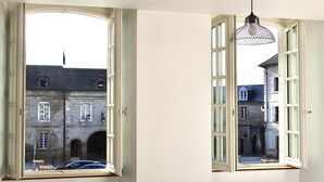 Una habitación con dos ventanas abiertas que dan a un edificio histórico de muros de piedra y numerosas ventanas.