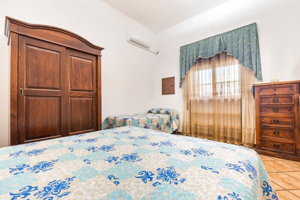 Un dormitorio con un amplio armario de madera, una cama con colcha estampada, una mesita de noche pequeña y una ventana con cortinas de encaje.