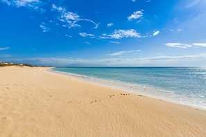 Uma praia com pegadas que levam à água, céu claro e mar calmo.