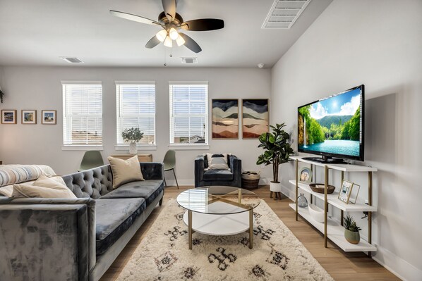 A modern living room with a grey sofa, a round glass coffee table, a TV on a white stand, and a ceiling fan. - Cozy Secluded Studio With Full-Kitchen, Washer And Dryer - The Works! (Austin)