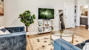 A modern living room with a blue sofa, a glass coffee table, a large flat-screen TV, a potted plant, and a white door leading to a kitchen. - Cozy Secluded Studio With Full-Kitchen, Washer And Dryer - The Works! (Austin)