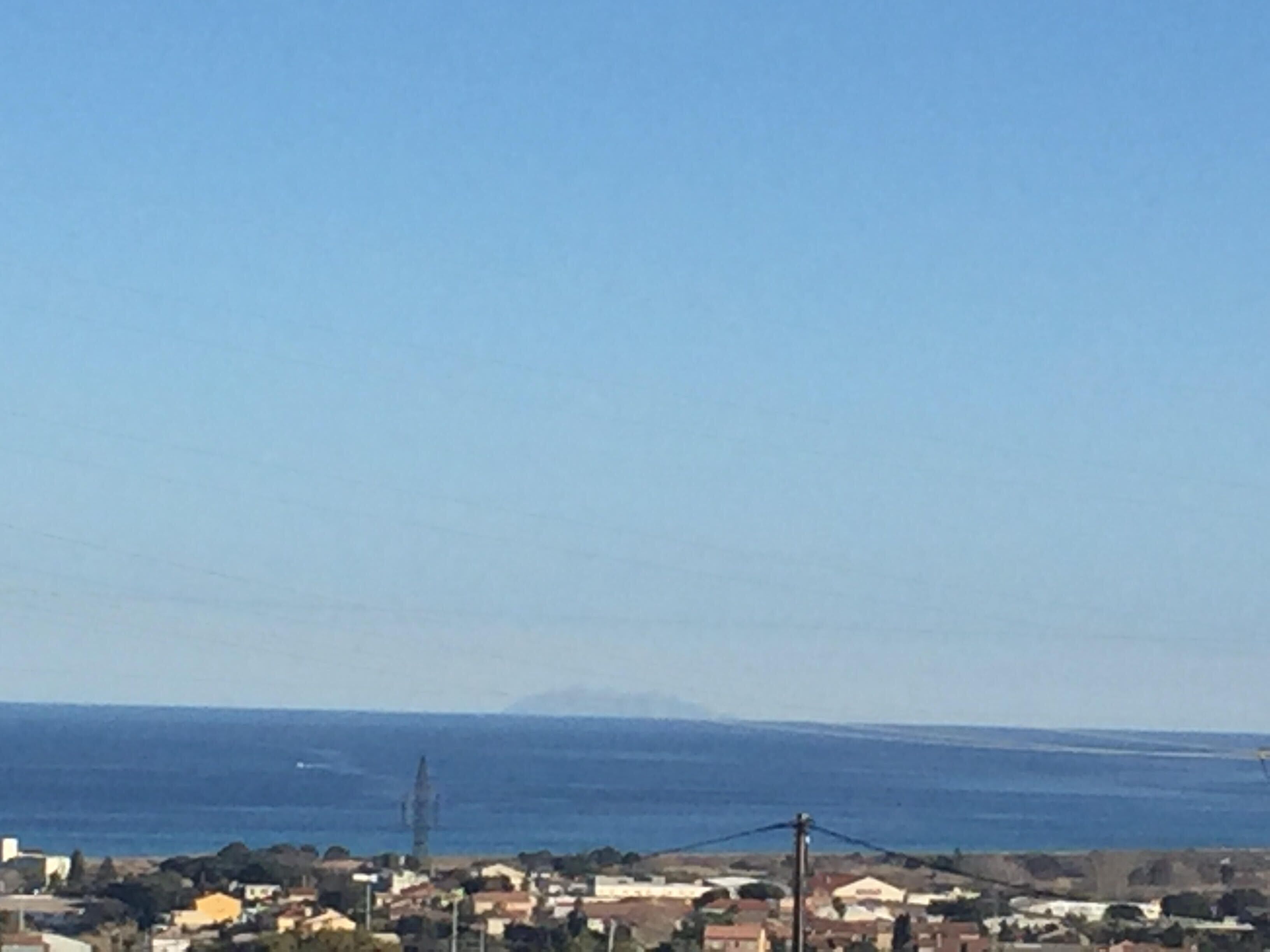 A coastal town with buildings along the shore, a clear sky, and a distant mountain.