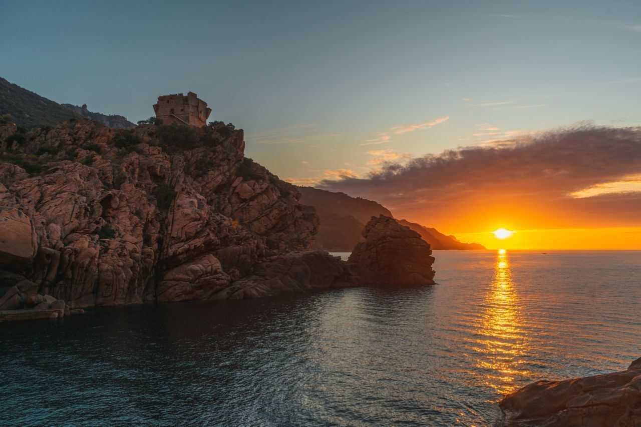 Un paisaje costero con un atardecer, acantilados rocosos y un pequeño edificio en la cima.