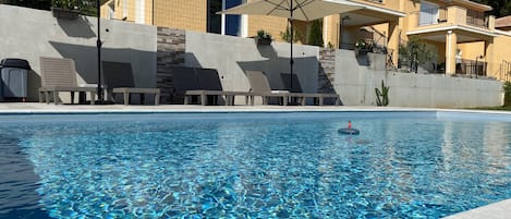 A swimming pool with clear water, lounge chairs, umbrellas, and a building with balconies.