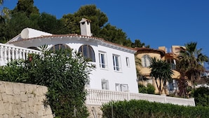 Una villa blanca con techo de tejas, múltiples ventanas y un muro de piedra al frente.