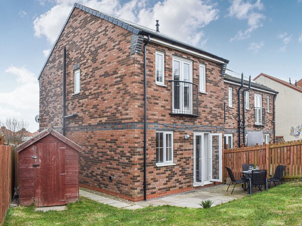 Une maison à deux étages, construite en briques, avec un toit en pente, un petit garage attenant et une cour arrière clôturée, aménagée avec une table et des chaises de patio.