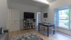A living room with a bookshelf, a TV, a dining table with chairs, and a window with a view of the outside.