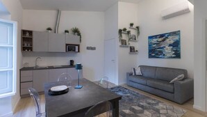 A modern living room with a grey sofa, a dining table with chairs, a kitchen area with cabinets and a microwave, and a painting on the wall.