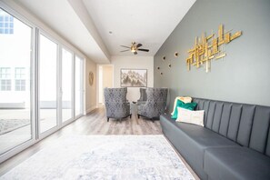 A modern living room with a grey sofa, two patterned armchairs, a ceiling fan, and a large window.