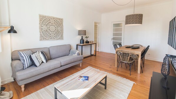 A modern living room with a grey sofa, wooden coffee table, and a dining area with a wooden table and wicker chairs.