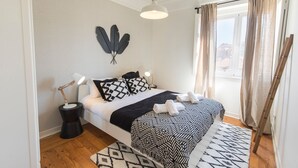 Un dormitorio moderno con una cama, dos mesitas de noche con lámparas, un adorno en la pared, una ventana con cortinas y una alfombra estampada.
