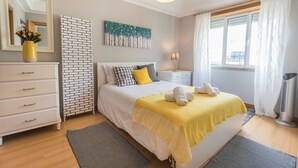 Een slaapkamer met een bed, een dressoir, een nachtkastje, een lamp, een schilderij en een raam met gordijnen.