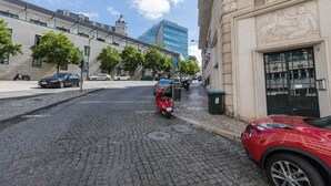 Een straat met kinderkopjes, geparkeerde auto's en een rode scooter.