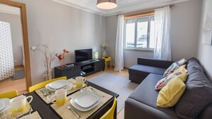 Een moderne woonkamer met een grijze bank, een eettafel gedekt voor twee personen, een televisie op een zwart meubelstuk en een raam met gordijnen.