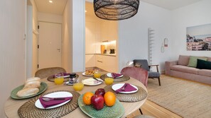 A modern living room with a dining area, featuring a round table set for breakfast with plates, glasses, and fruits.