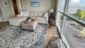 A modern living room with a sectional sofa, a coffee table with a plant, a large window with a view of the sea, and a patterned rug.