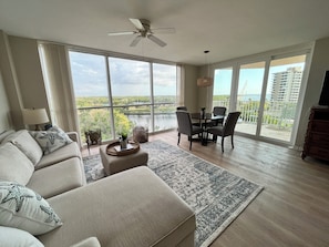 Relax in this serene space with a panoramic view of the water and surrounding greenery, featuring a comfortable sofa, dining table, and mounted TV.