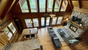 A cabin interior with a wooden floor, a stone fireplace, a sofa, a dining table with chairs, and large windows offering a view of the outdoors. - Beautiful secluded cabin - sleeps 12, fire pit, fishing pond, grill, game room (russell springs)