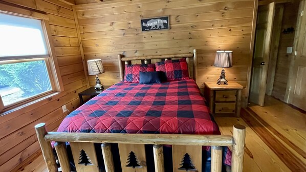 Une chambre dans une cabane en bois, avec un lit, deux tables de chevet, une petite table, une fenêtre et un panneau accroché au mur.
