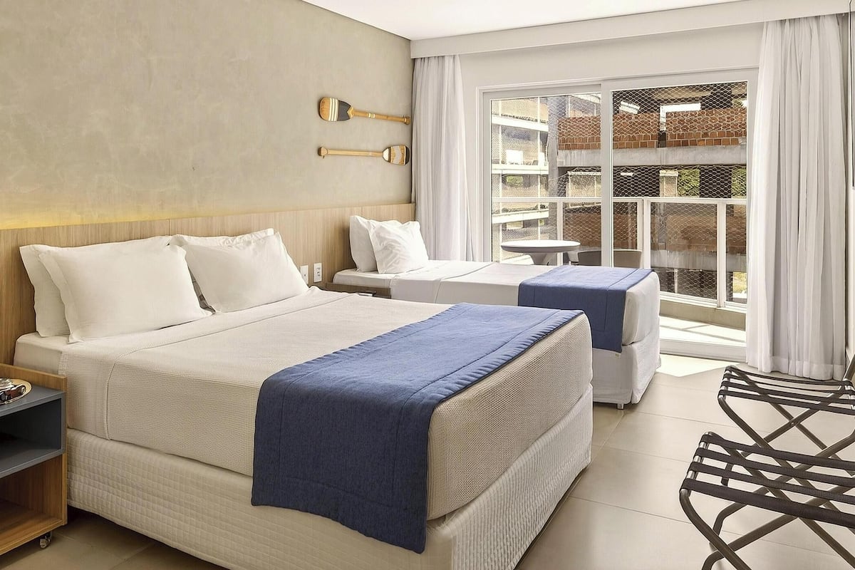 Een hotelkamer met een groot bed, een kleiner bed, een bureau en een stoel. Er is een raam met uitzicht op gebouwen.