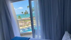 A hotel room with a bed, a view of a pool and beach, and a balcony with curtains.