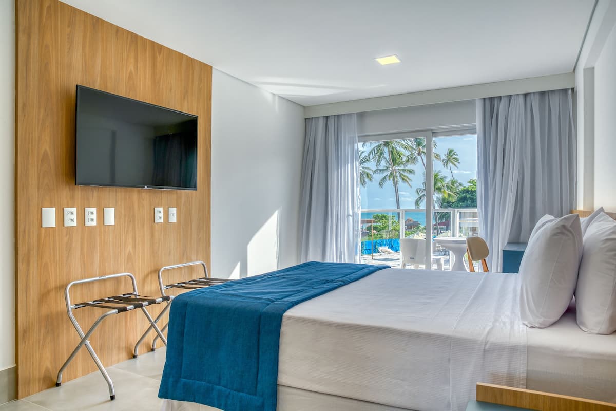 Een hotelkamer met een bed, een televisie bevestigd aan een houten wand, een balkon met uitzicht op palmbomen en een stoel bij het raam.