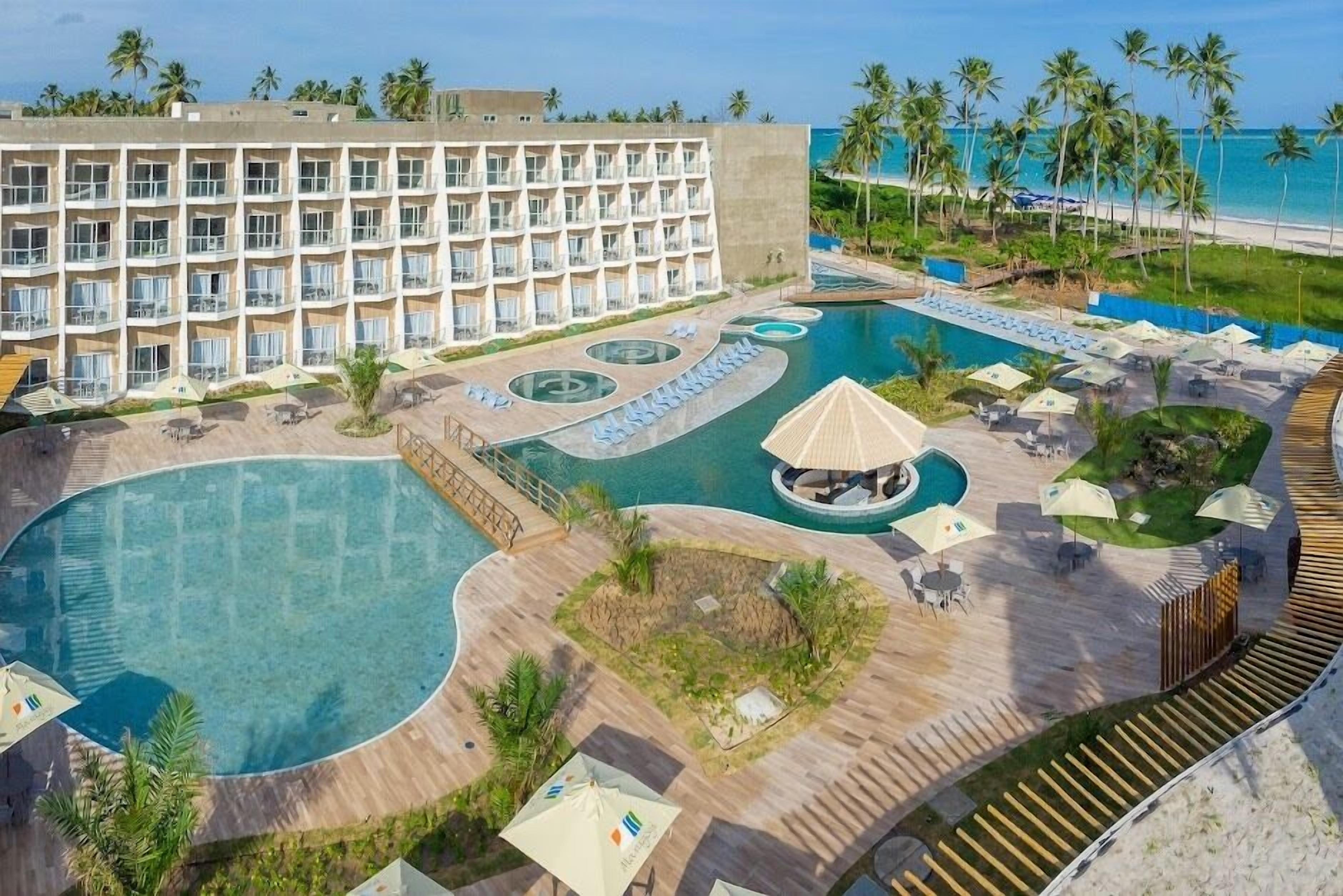 Un hotel con varios balcones, una gran piscina central y vista a la playa.