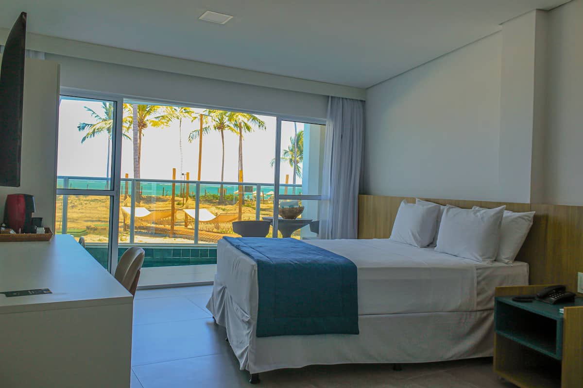 Een hotelkamer met een bed, een bureau en uitzicht op palmbomen en het strand.