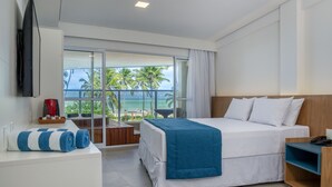 A modern hotel room with a large bed, a TV mounted on the wall, a balcony with a view of palm trees and the ocean, and a desk with a coffee maker.